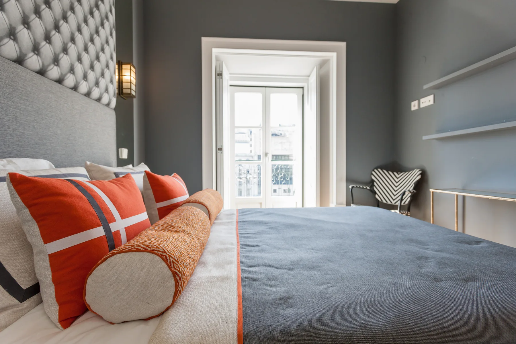 Dormitorio con una cama, cojines coloridos y una silla junto a la ventana.