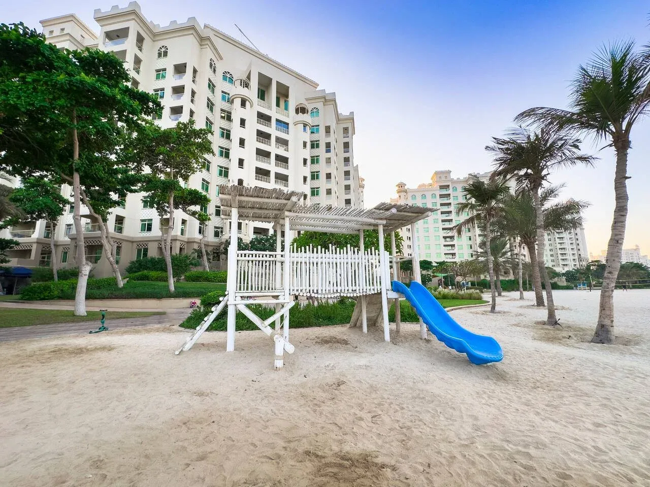 A sandy playground with a blue slide sits among palm trees and tall buildings.