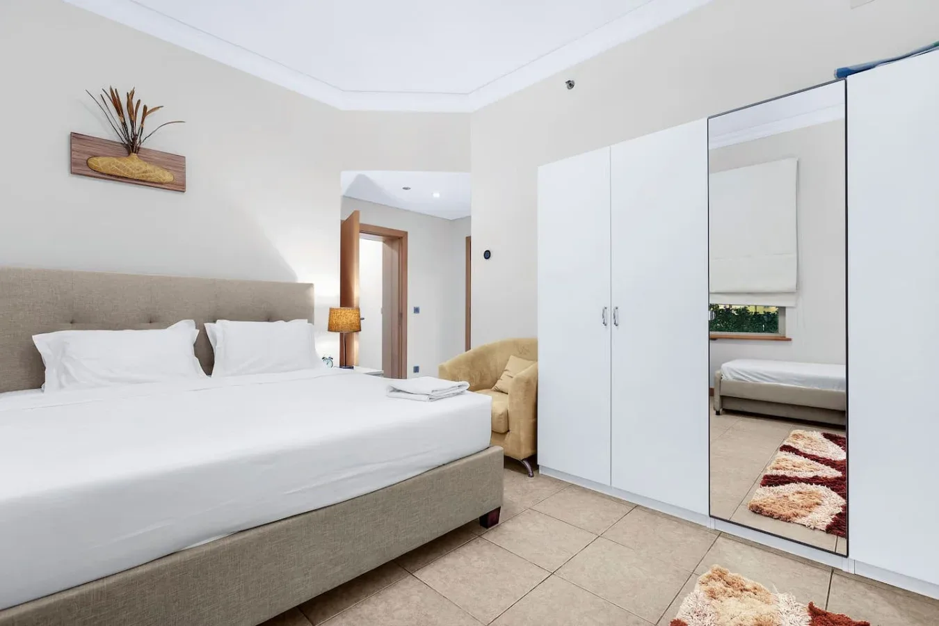 A bright bedroom with a large bed, white wardrobes, and a mirrored closet door.