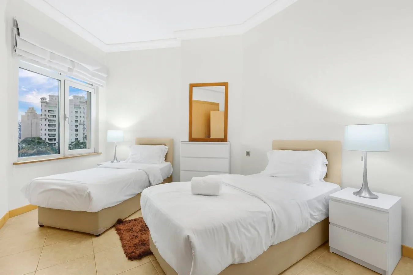 A bright bedroom with two beds and a window overlooking nearby buildings.