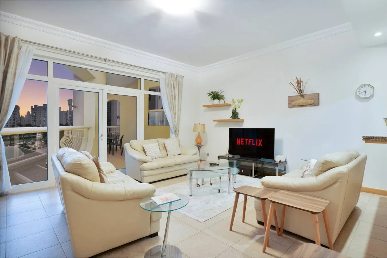 Una sala luminosa con asientos color crema, una mesa de centro de vidrio y un televisor.