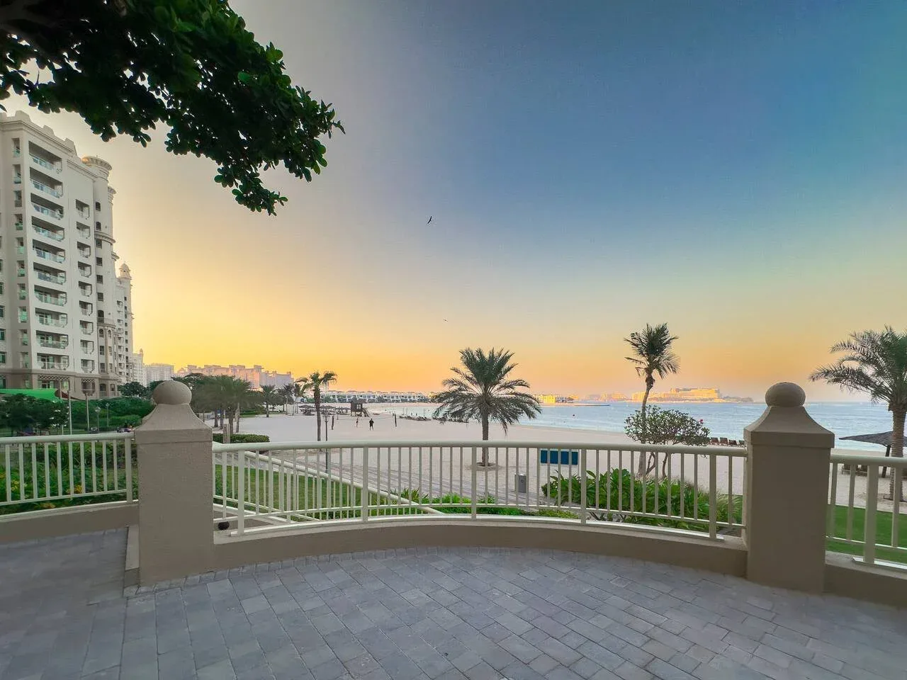 A seaside promenade overlooks the water at sunset, with palm trees along the shore.