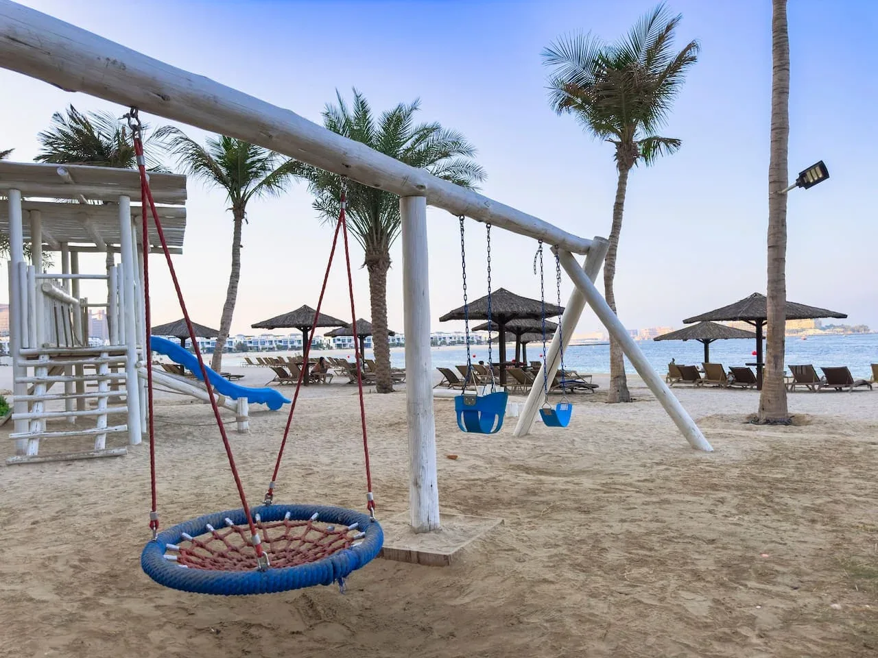 A sandy beach play area with swings and palm trees beside the water.
