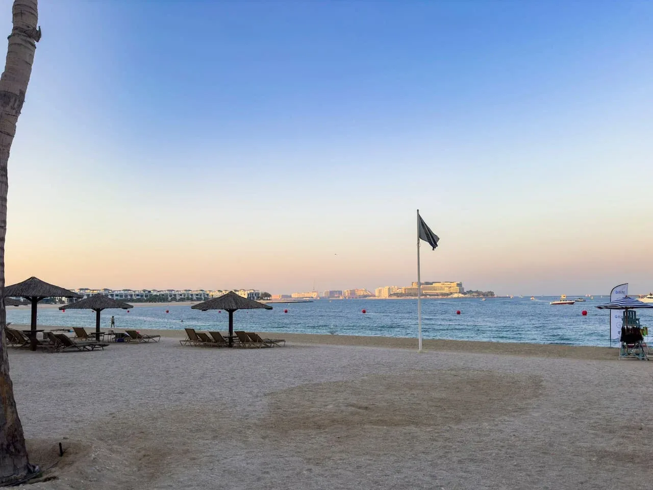 A sandy beach at dusk with the sea and a flag in the distance.