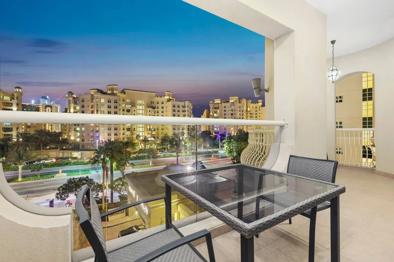 A balcony dining area overlooks illuminated buildings and a road at dusk.