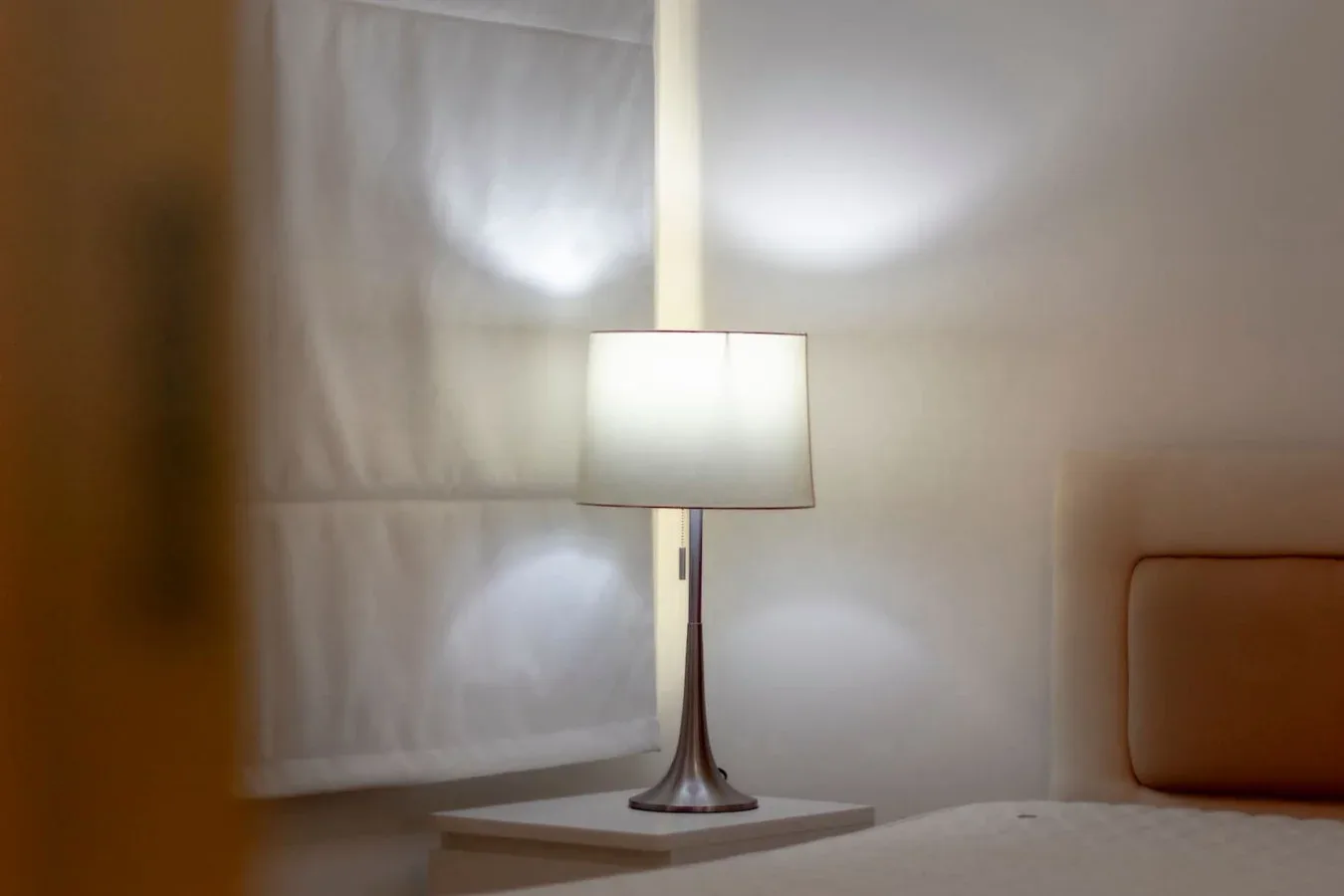 A lit table lamp stands beside a window with a pale shade.