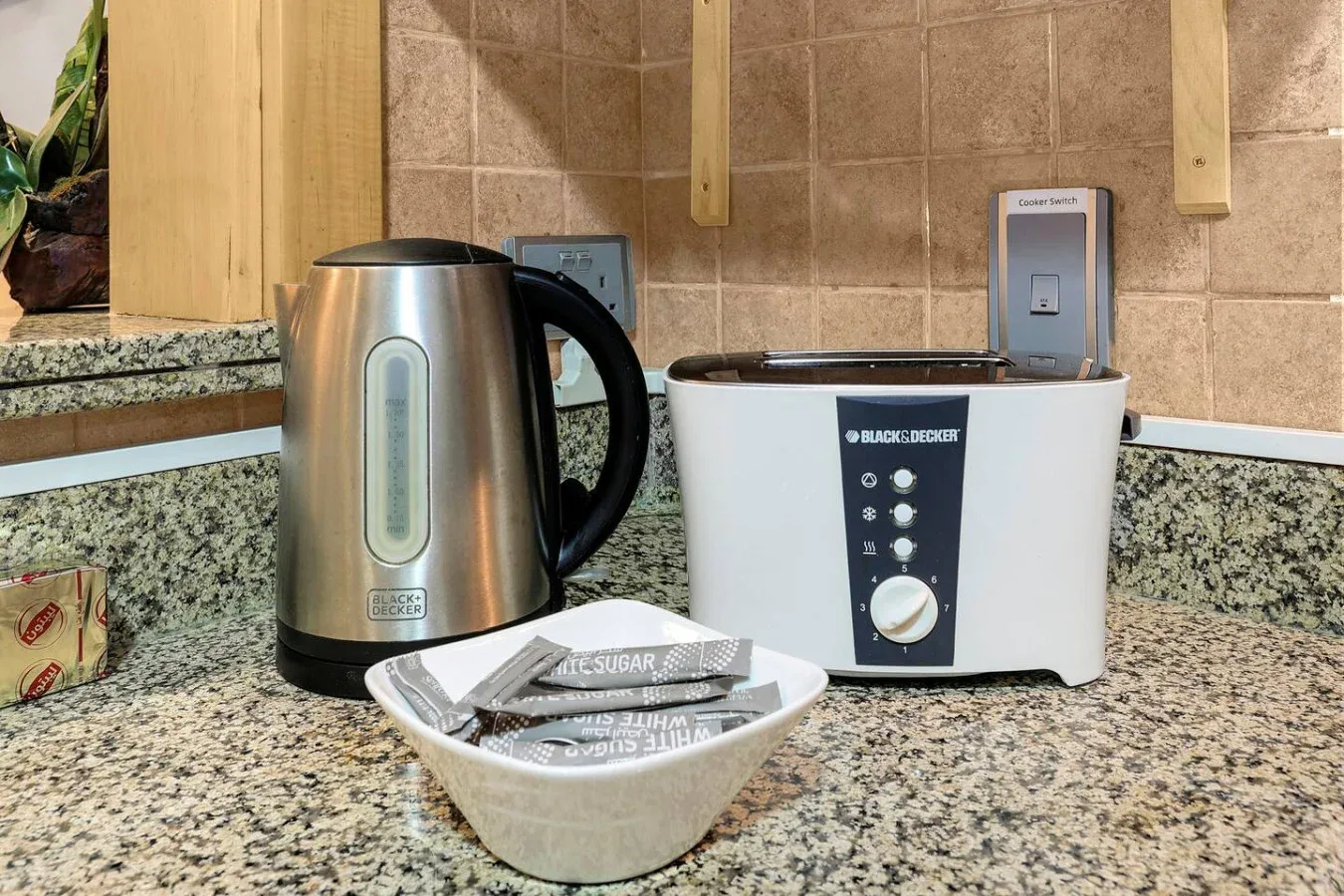 A kitchen counter holds a kettle, a toaster, and a dish of wrapped utensils.