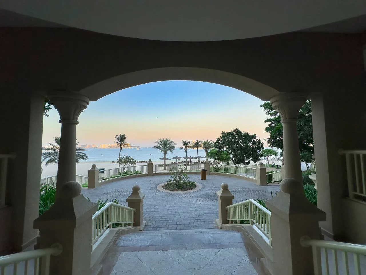 A covered terrace opens onto a paved courtyard with palm trees and water beyond.