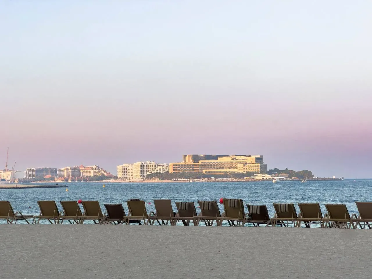 Seaside view with lounge seating along the shore and buildings across the water.
