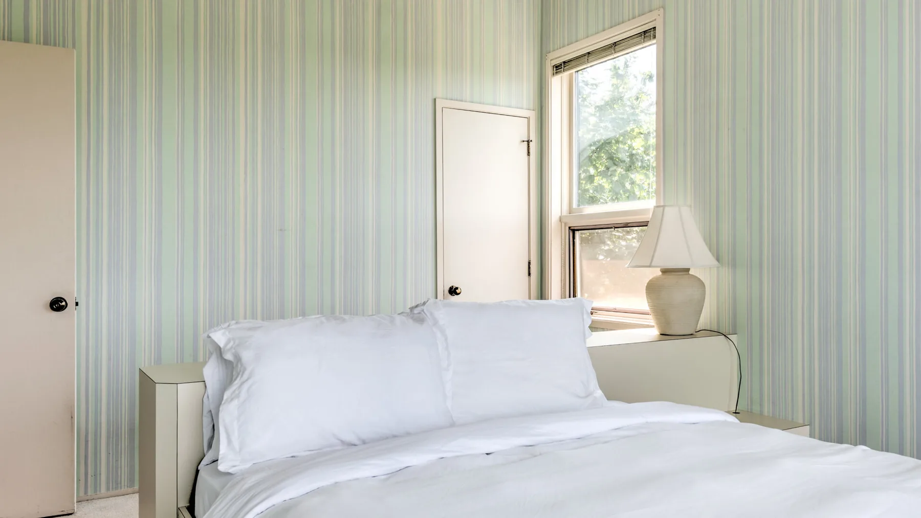 Un dormitorio con ropa de cama blanca, papel tapiz a rayas y una ventana.