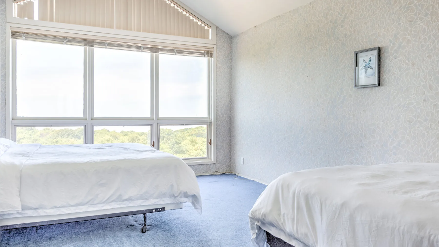 Un dormitorio luminoso con dos camas, ventanas grandes y un pequeño cuadro enmarcado.