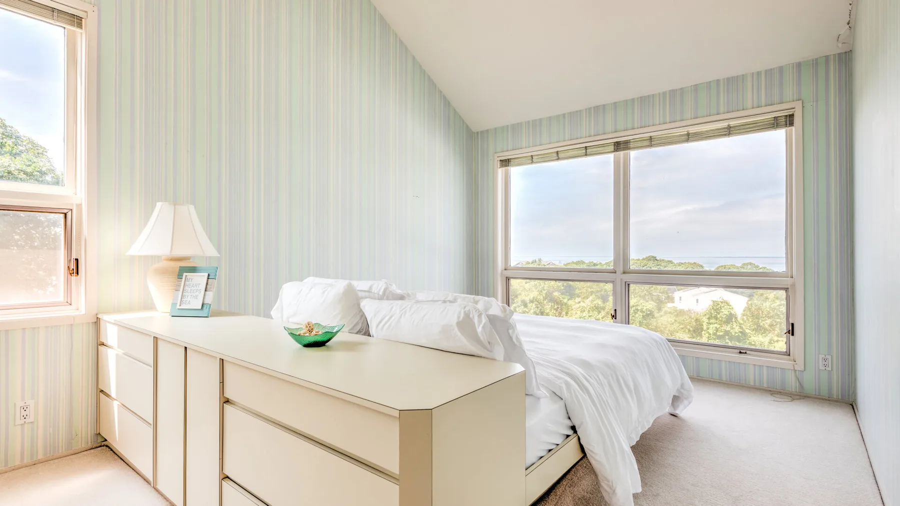 Un dormitorio luminoso con una cama, ropa de cama blanca y papel pintado a rayas.