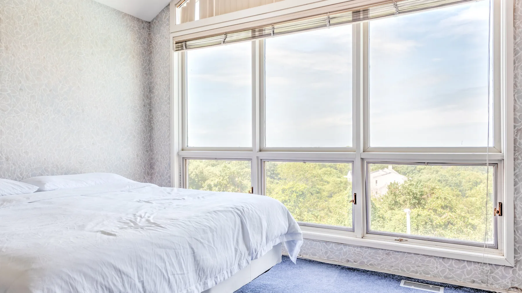 Un dormitorio con una cama junto a grandes ventanas con vista a árboles verdes.
