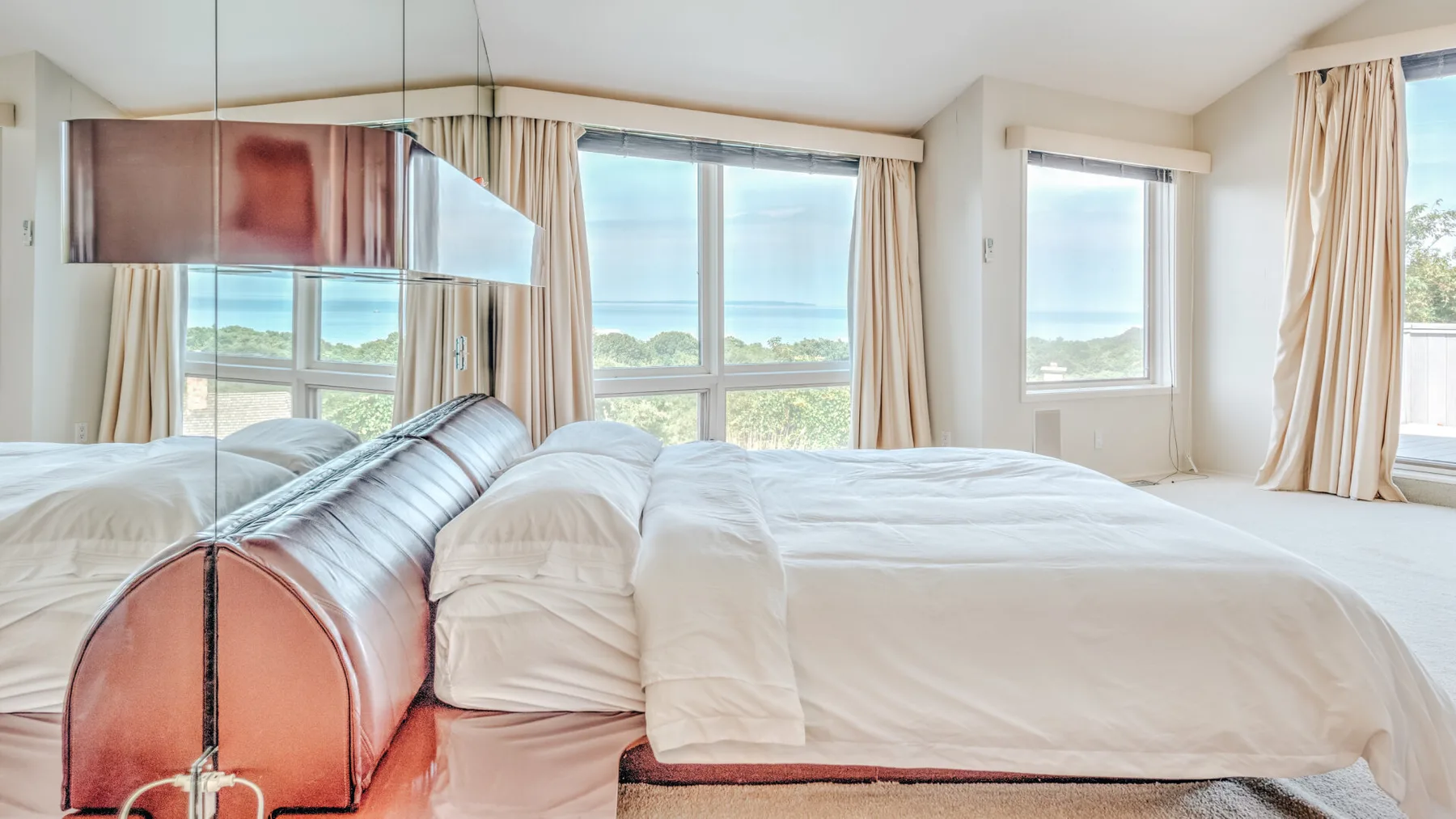 Un dormitorio con ropa de cama blanca, ventanas grandes y un panel reflectante junto a la cama.