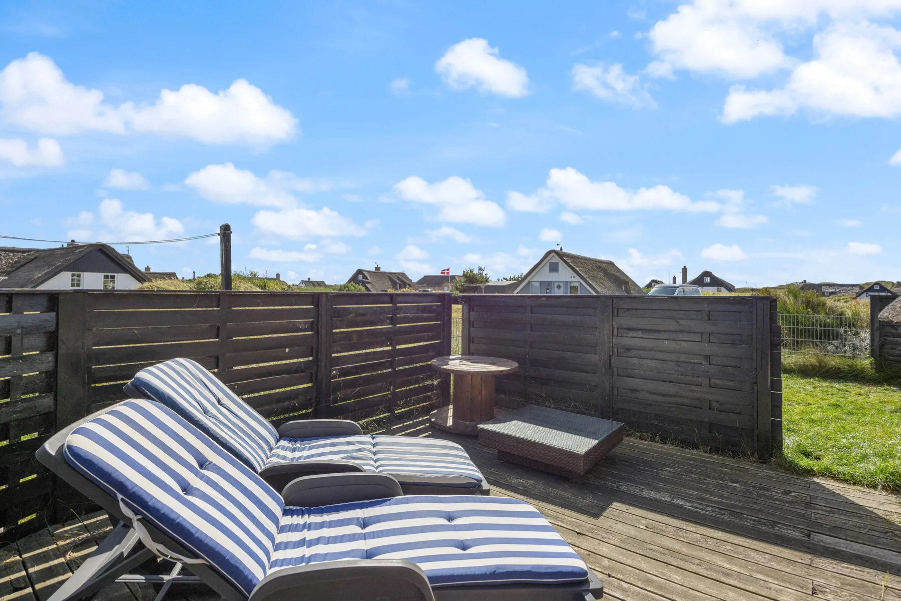 Outdoor deck seating with striped cushions, a wooden privacy fence, and sunny skies.