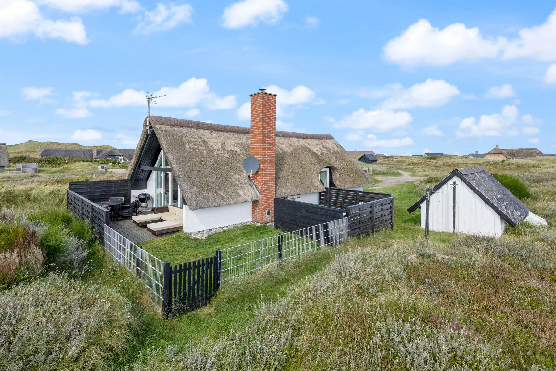 A thatched-roof house sits among grassy dunes beneath a blue sky.