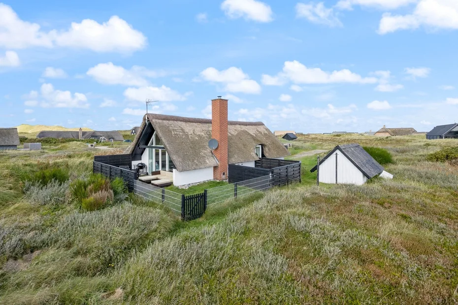 A small house sits among grassy dunes beneath a partly cloudy sky.