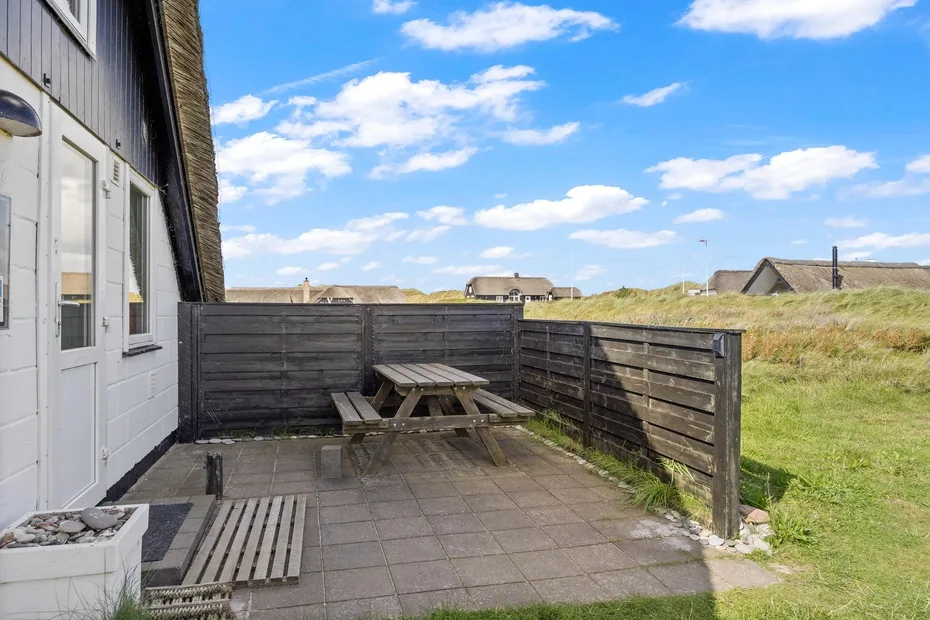 A paved outdoor patio with a wooden picnic table sits beside a fenced grassy area.