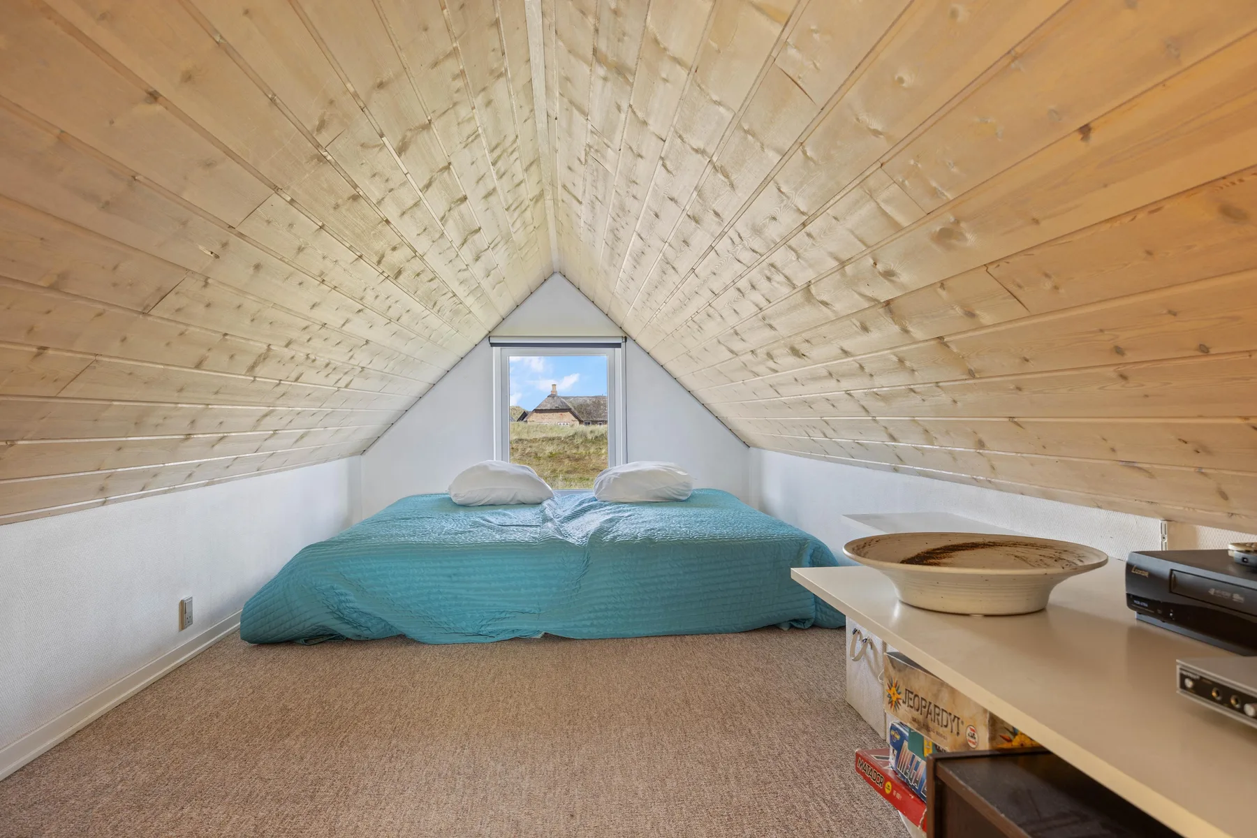A low bed sits beneath a sloped wooden ceiling, with a small window behind it.