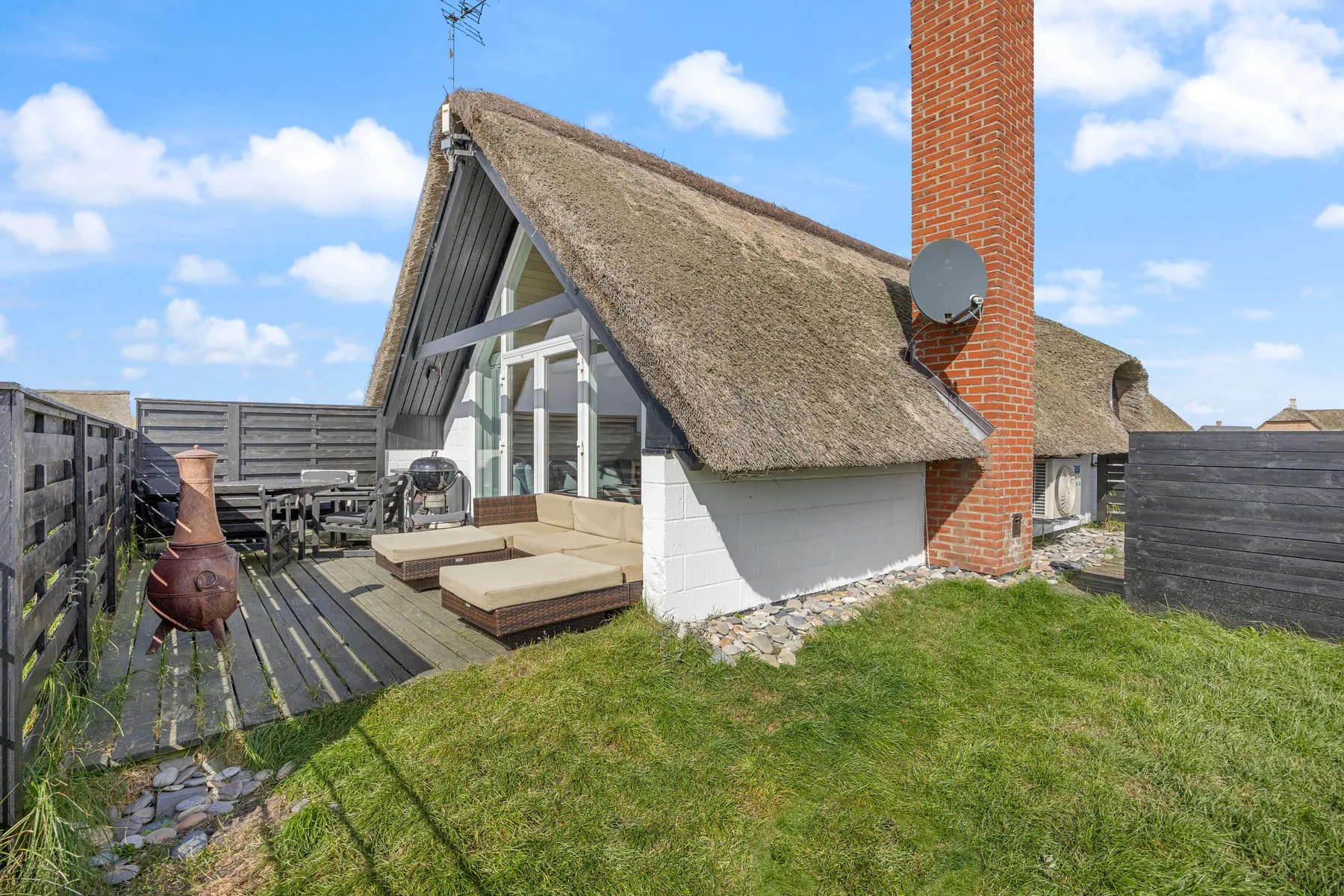 A thatched-roof cottage beside a wooden deck and grassy yard.