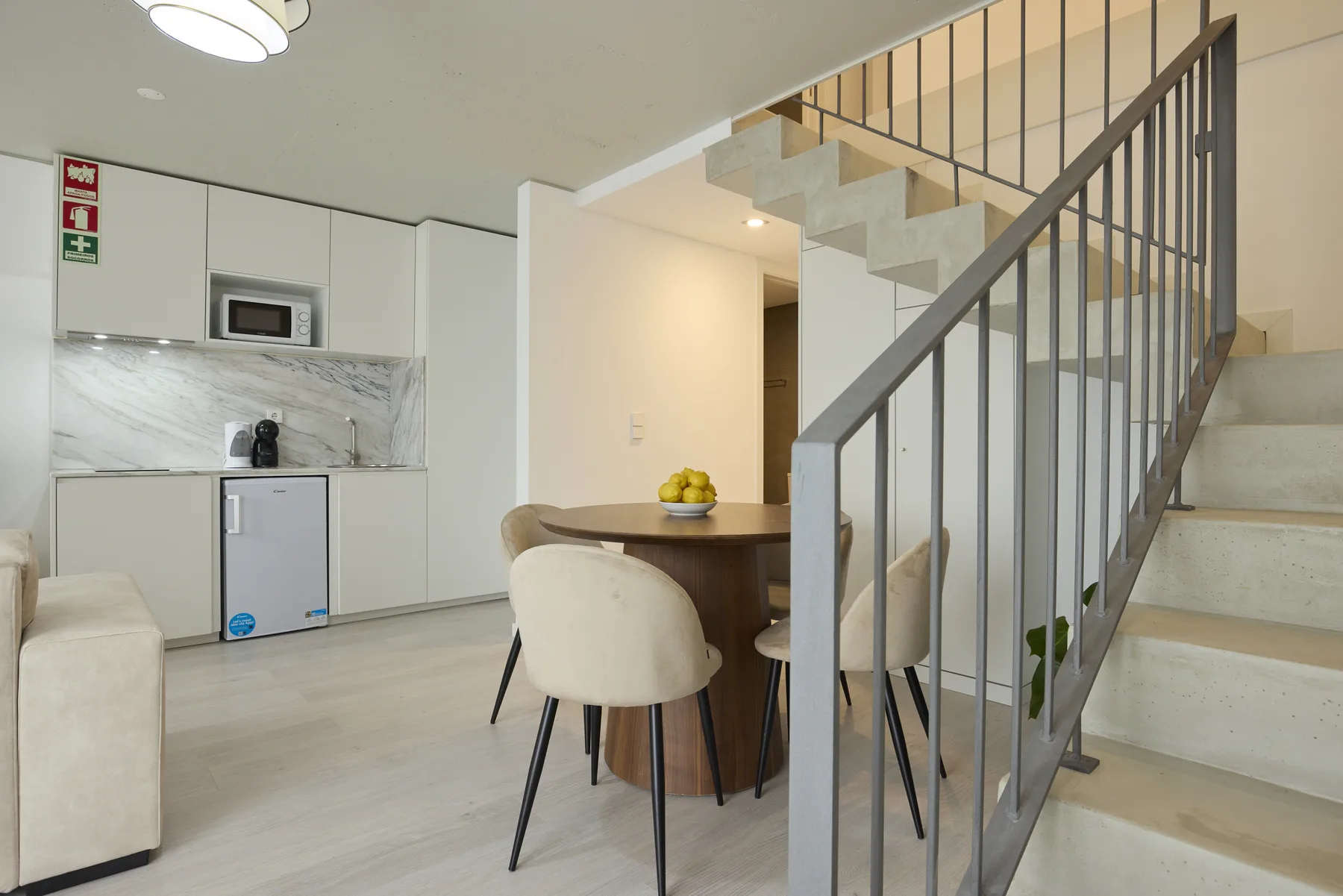 A dining area with a round table sits beside a staircase and compact kitchen.
