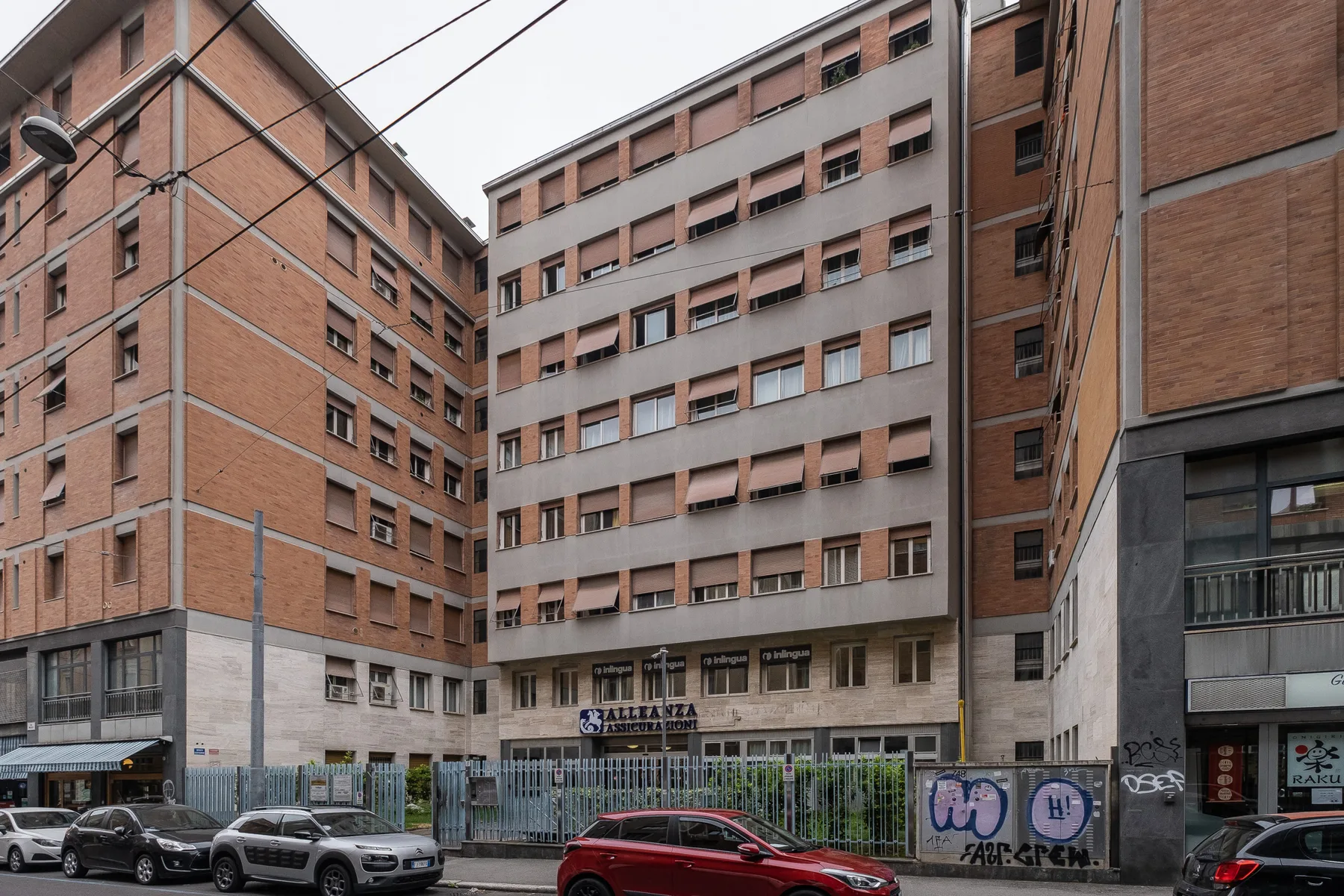 Una calle urbana bordeada de edificios de varias plantas, coches aparcados y una acera cercada.