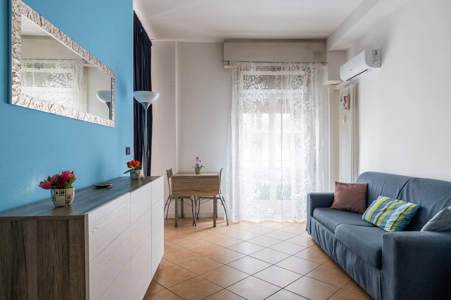 Una sala luminosa con un sofá azul y una pequeña mesa de comedor junto a la ventana.