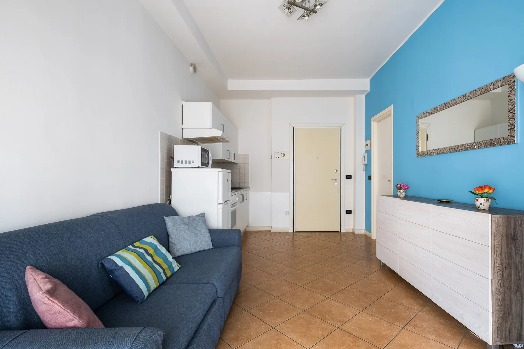 Una sala de estar con asientos, una pequeña cocina y una cómoda blanca junto a una pared azul.