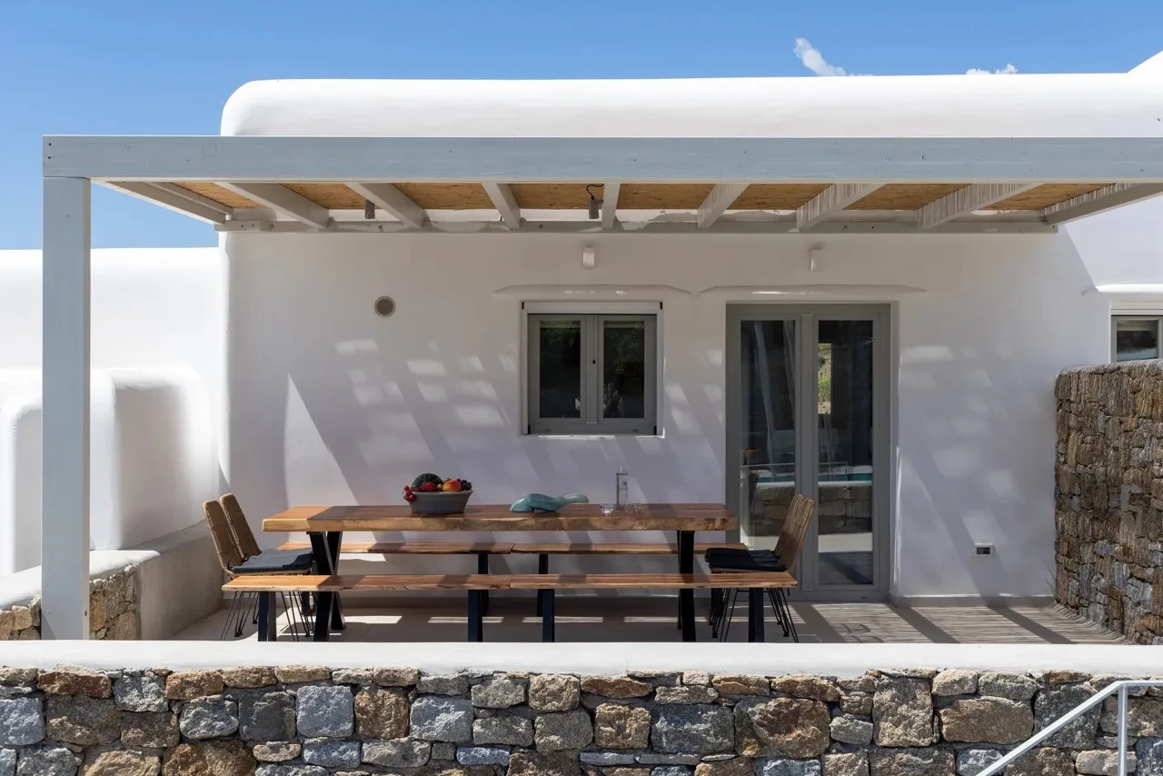 A shaded outdoor dining area with a wooden table beside a white building.