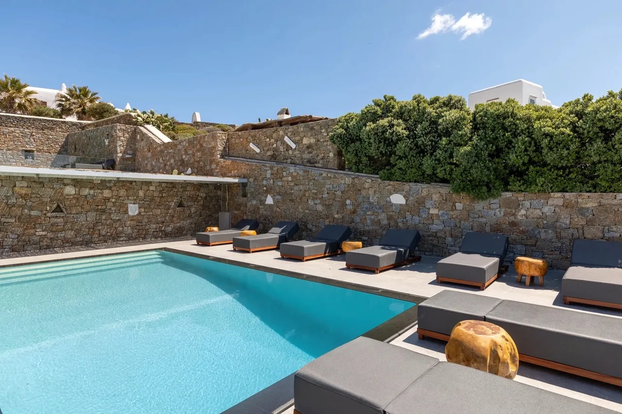 An outdoor swimming pool is surrounded by dark lounge seating and stone walls.
