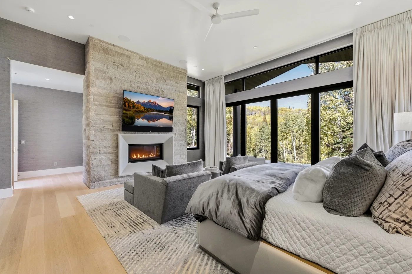A bright bedroom with a bed, seating area, and large windows overlooking trees.