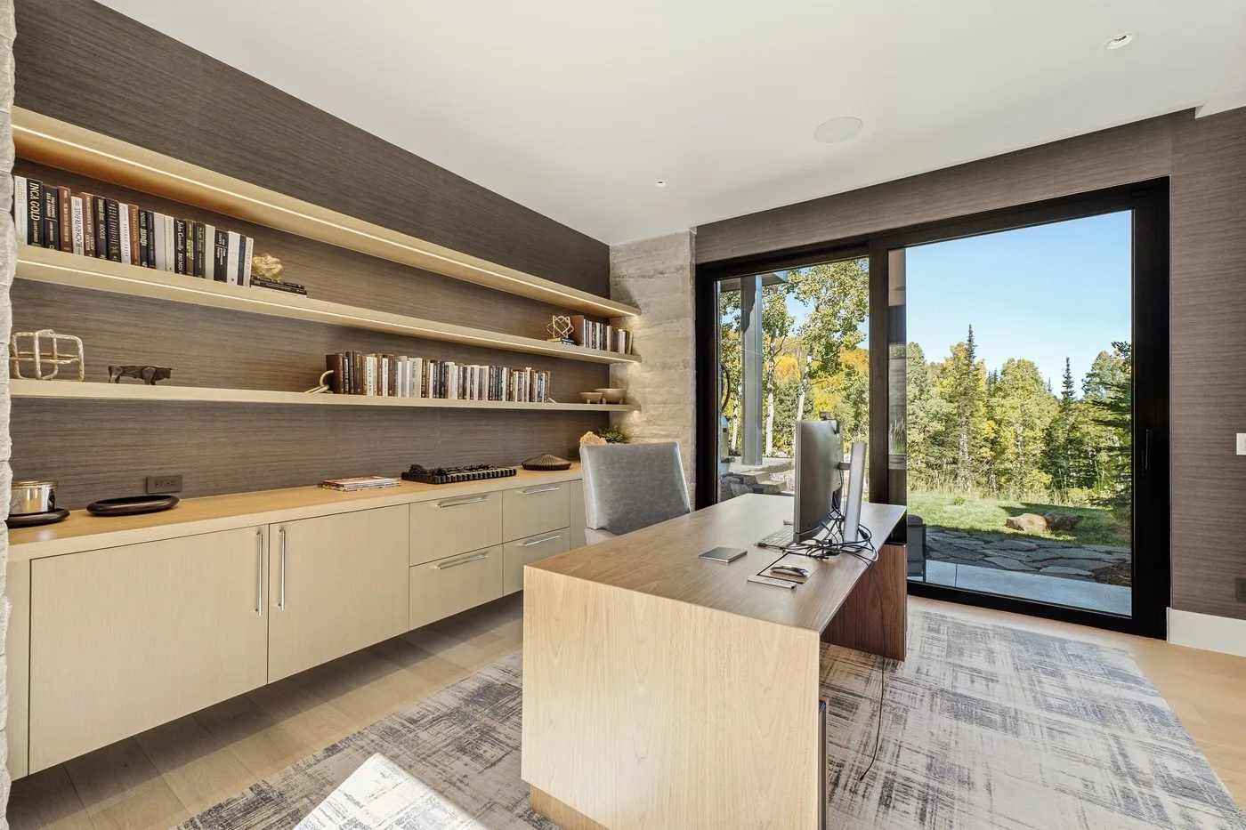 A modern office with a desk, shelving, and a large window overlooking trees.
