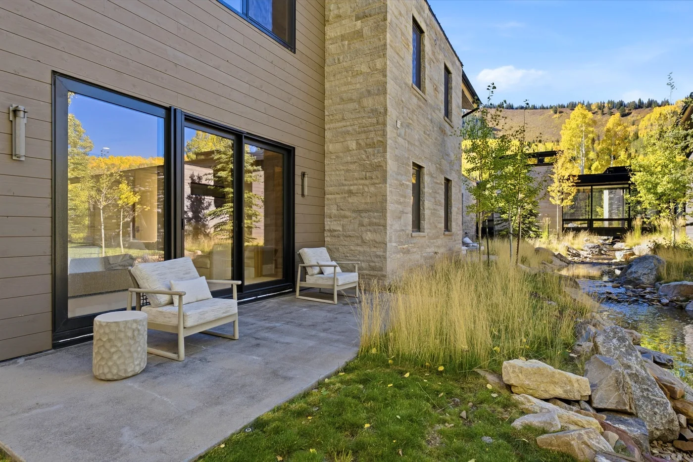 A modern building opens onto a patio with outdoor seating beside a landscaped stream.