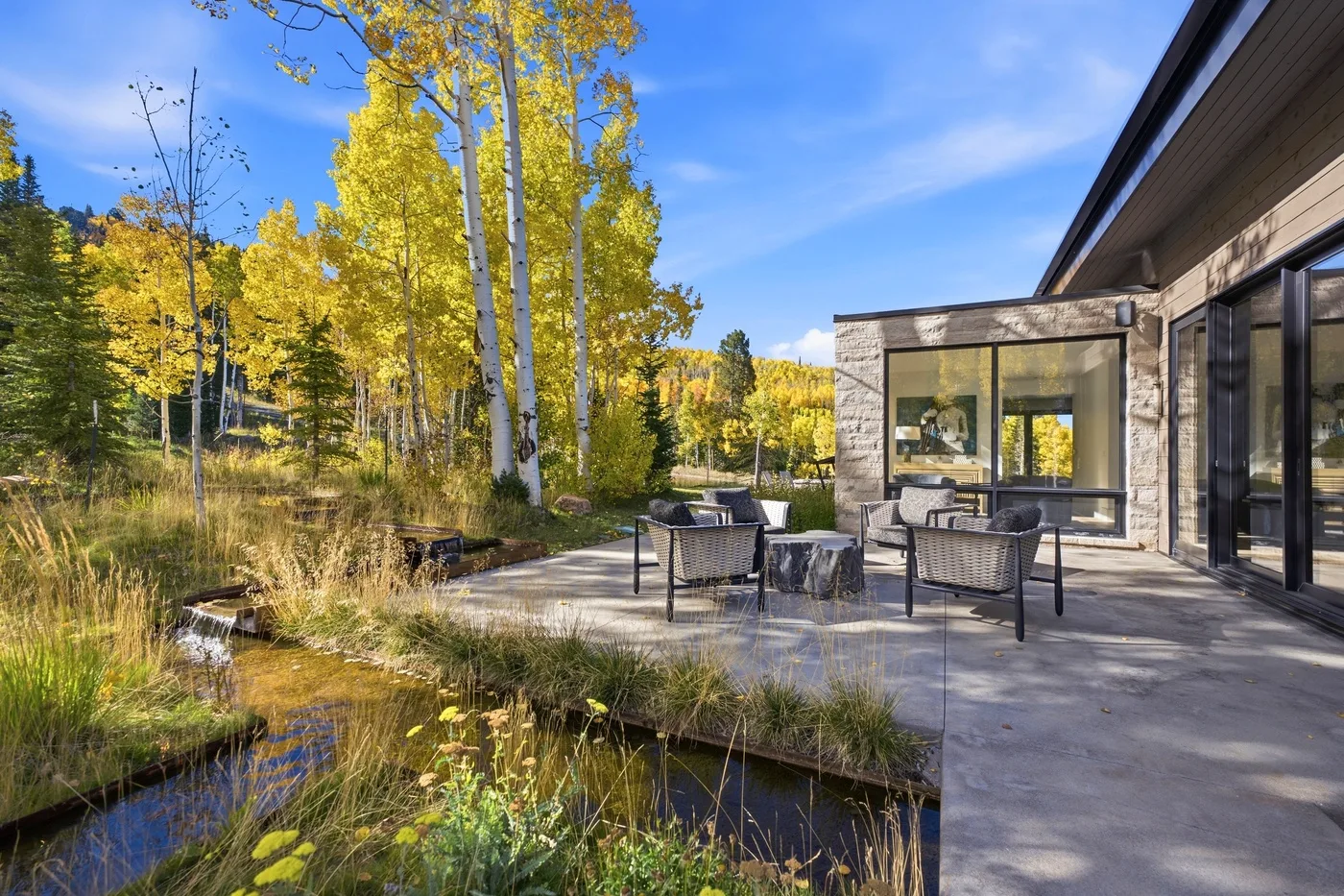 An outdoor patio beside a modern building overlooks trees and a landscaped pond.