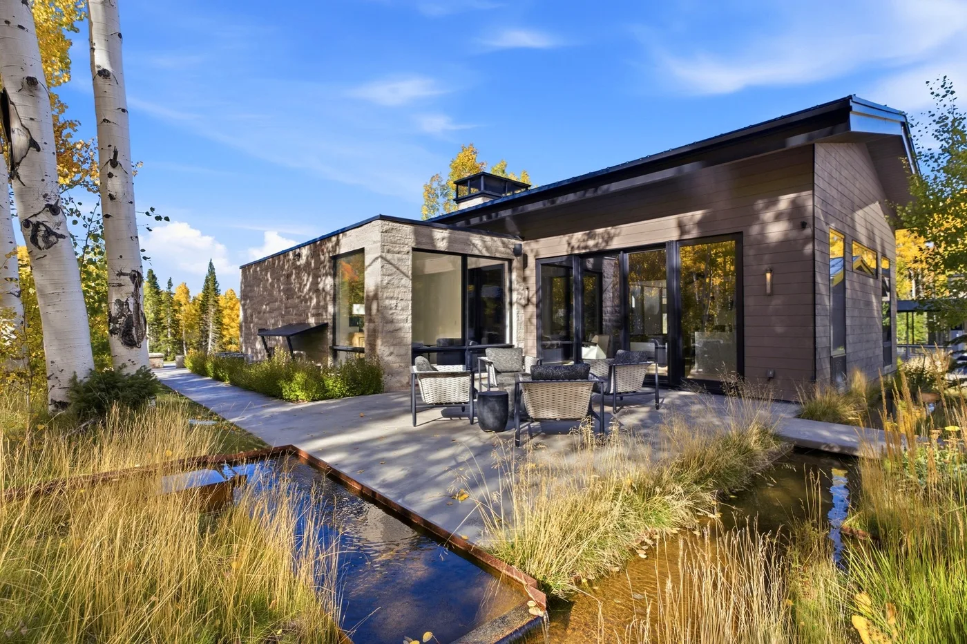 A modern house opens onto a paved patio with outdoor seating beside a pond.