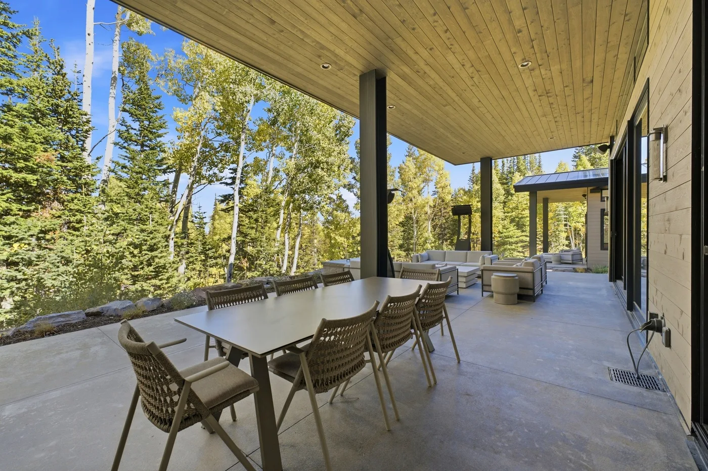 A covered outdoor patio has a dining table and seating beside a wooded view.