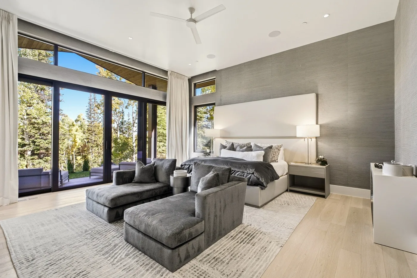 A bright bedroom with a large bed, gray seating, and floor-to-ceiling windows.