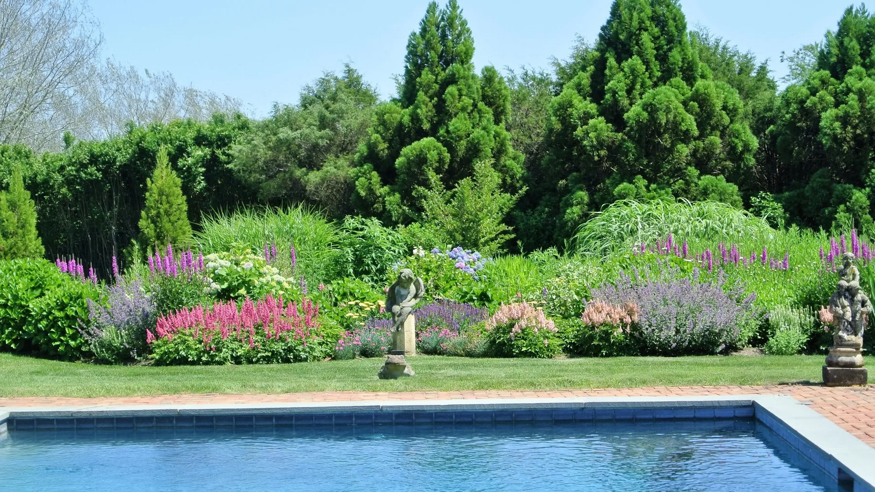 Una piscina bordea un jardín frondoso con plantas en flor y estatuas.