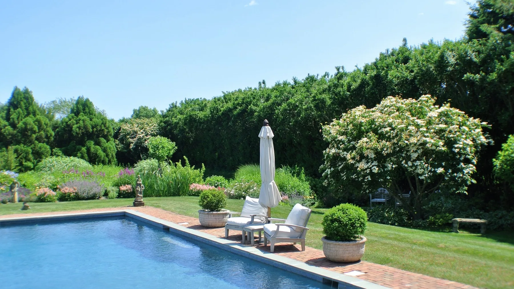 Una piscina al aire libre junto a un patio pavimentado con asientos y vegetación.