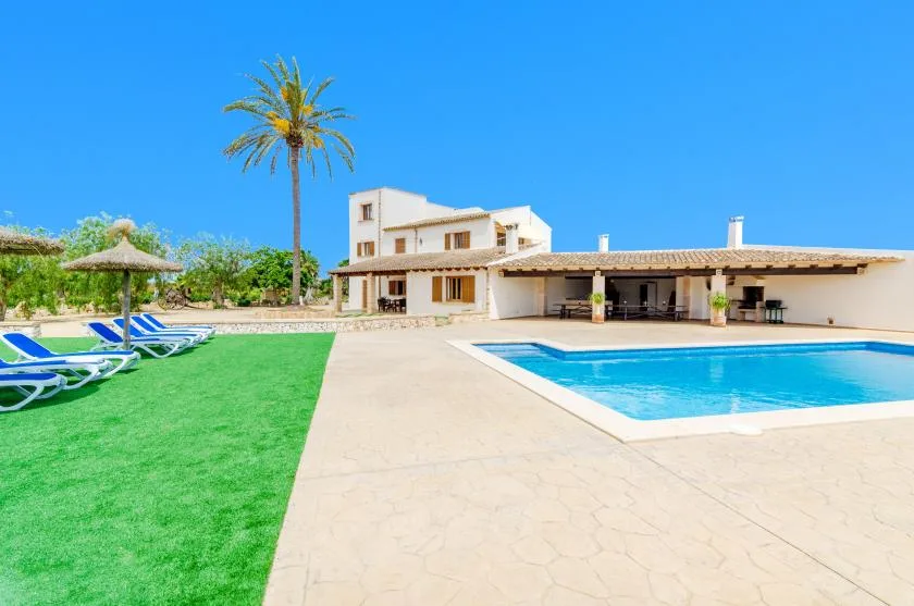 An outdoor swimming pool beside a large house, with green lawn and lounge seating.