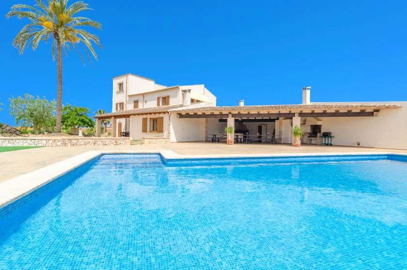 A swimming pool sits in front of a large house with a covered patio.