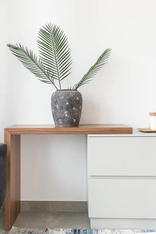 Una planta frondosa en una maceta oscura está sobre una encimera de madera junto a cajones blancos.