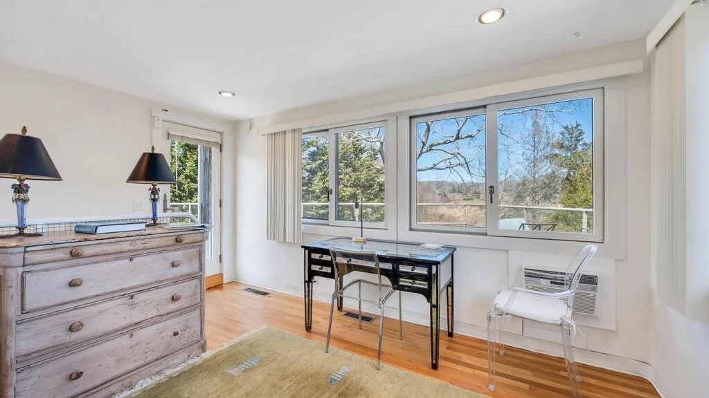 Una habitación luminosa con un escritorio y grandes ventanas que dan a los árboles.