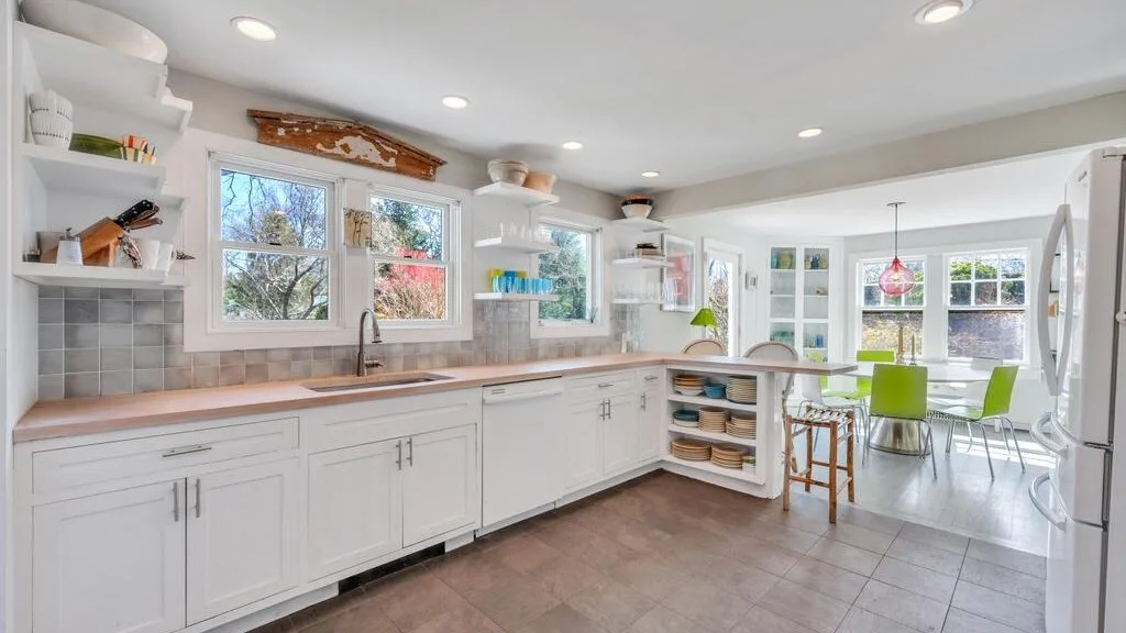 Una cocina luminosa con gabinetes blancos, encimeras de madera y un comedor.