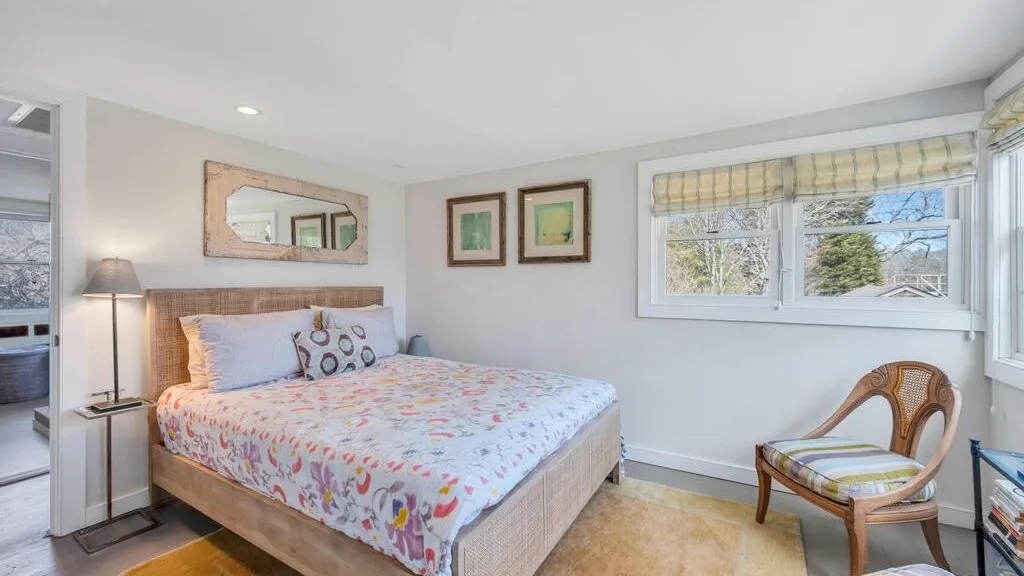 Un dormitorio luminoso con una cama, una lámpara de noche y ventanas con vista a los árboles.