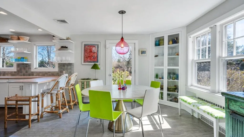 Un comedor luminoso con una mesa y asientos coloridos junto a grandes ventanas.