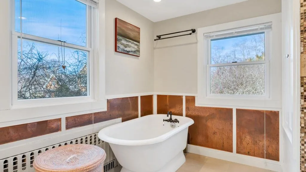 Un baño luminoso con una bañera exenta bajo dos ventanas.