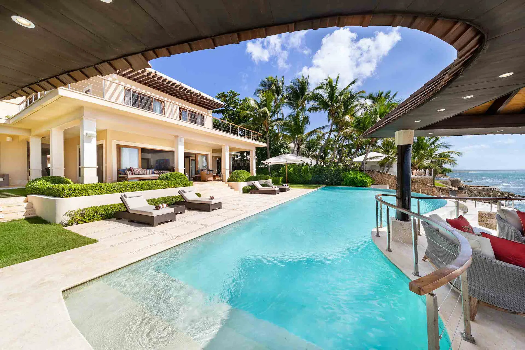 A coastal poolside patio beside a modern house, with palm trees and the sea beyond.