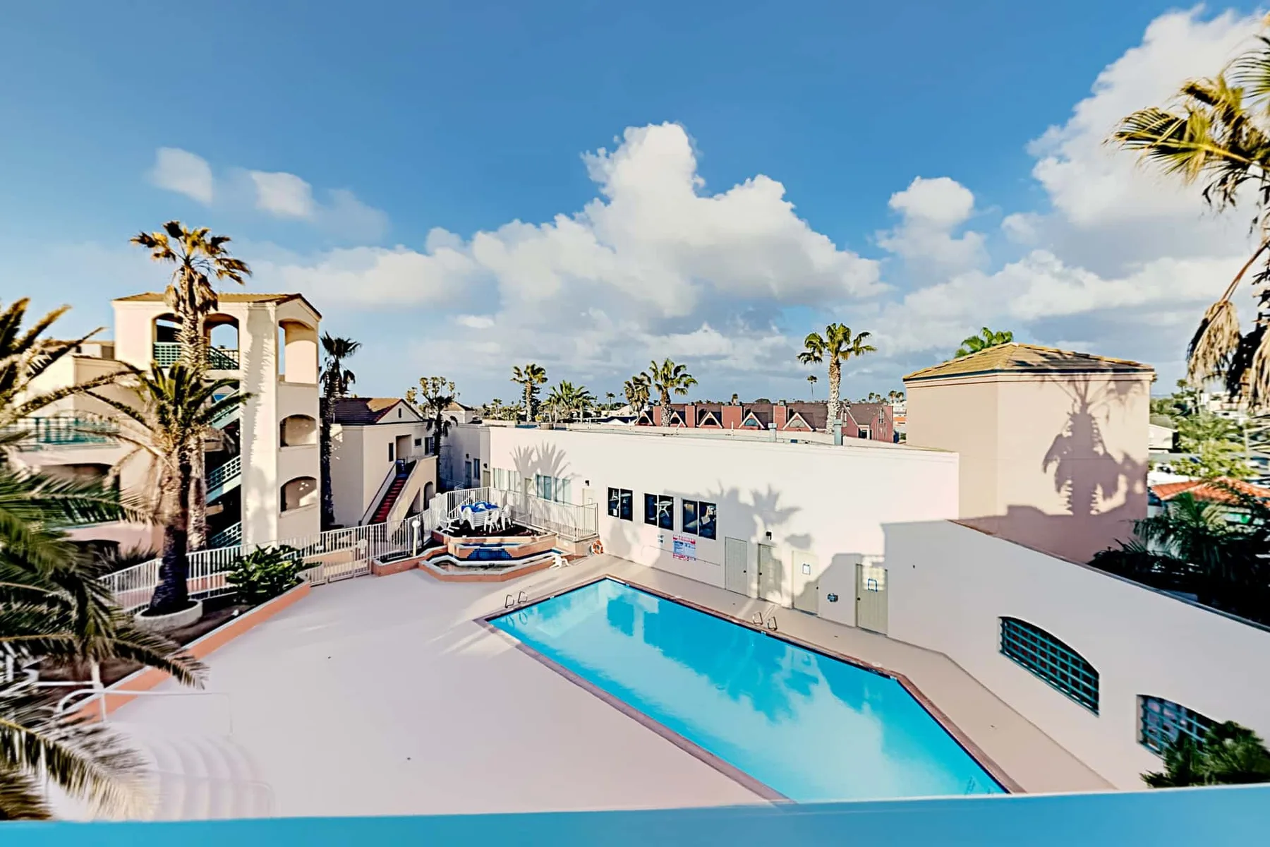 Una piscina en la azotea está junto a un edificio claro, con palmeras y cielo azul a la vista.