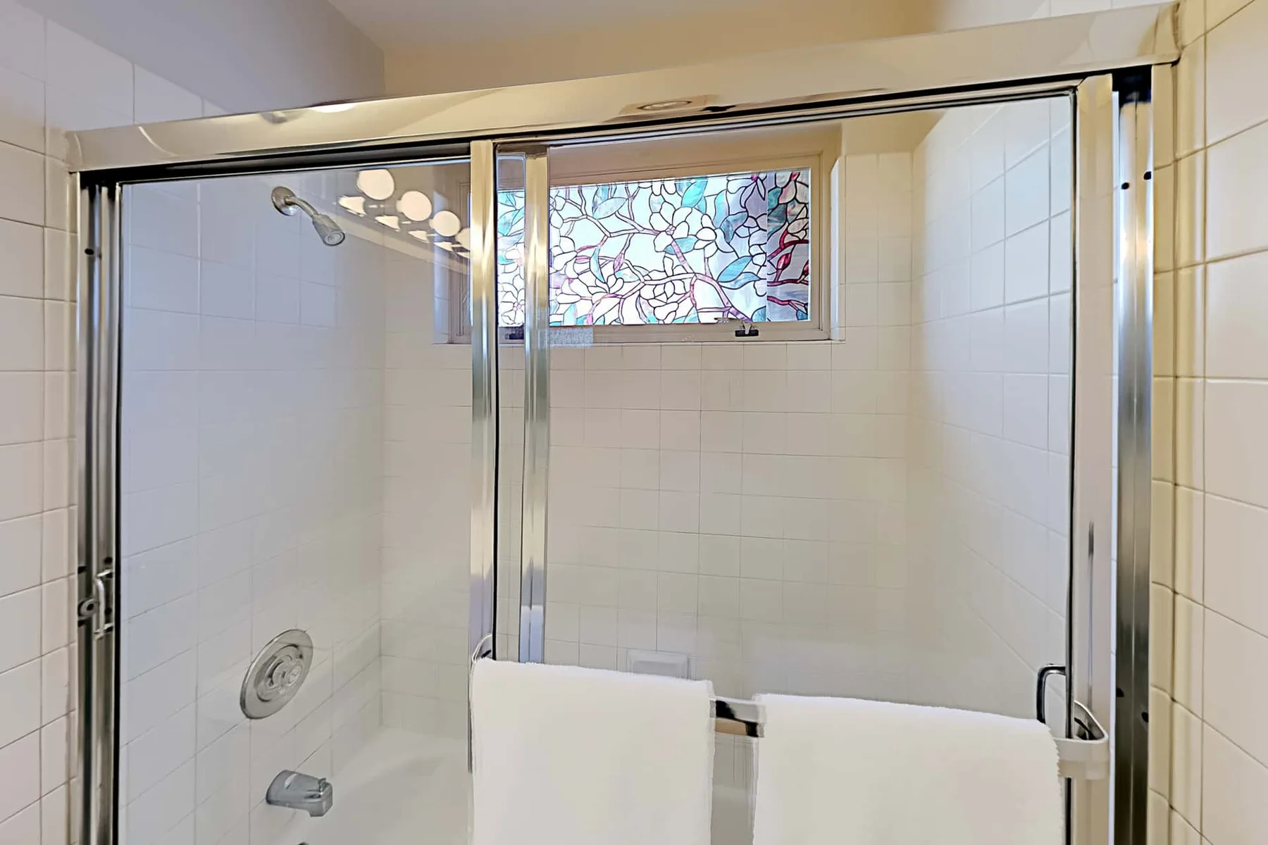 Una ducha con azulejos, una ventana decorada y toallas colgadas sobre el vidrio.